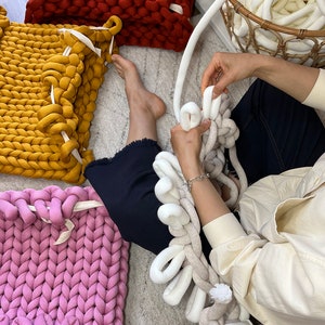 May include: A person is making a chunky knit blanket with white yarn. The blanket is made of large, thick loops of yarn. There are other chunky knit blankets in the background, in red, yellow, and pink.