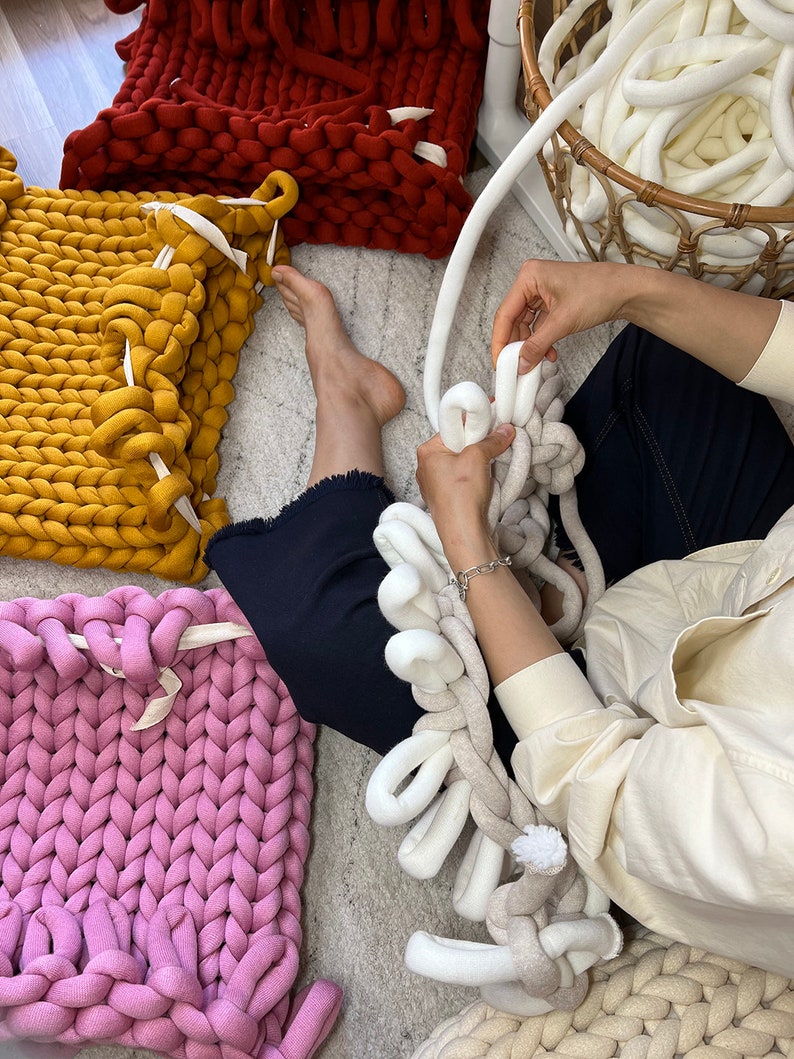 May include: A person is making a chunky knit blanket with white yarn. The blanket is made of large, thick loops of yarn. There are other chunky knit blankets in the background, in red, yellow, and pink.