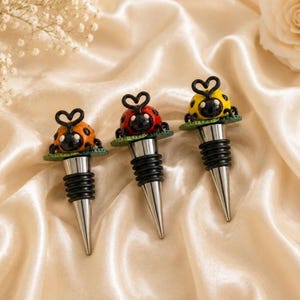 May include: Three decorative bottle stoppers with ladybug designs. Each stopper features a ladybug with a black head and a body in orange, red, or yellow. The stoppers have a silver cone and a black rubber seal. The ladybugs sit on a green base.
