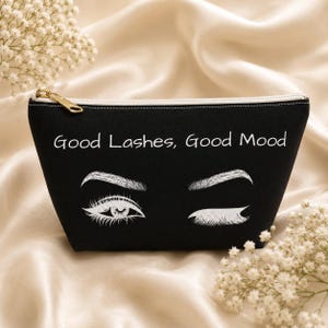 Könnte beinhalten: Eine schwarze Reißverschlusstasche mit dem weißen Aufdruck Good Lashes, Good Mood. Unter dem Text befindet sich eine weiße grafische Illustration, die ein offenes Auge mit langen Wimpern und ein zwinkerndes Auge mit Augenbraue darstellt.