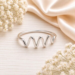 May include: A polished silver ring featuring a continuous, smooth, undulating wave pattern that mimics a heartbeat rhythm. The band is crafted from a single piece of reflective metal, creating a clean, minimalist, and fluid design that encircles the finger.