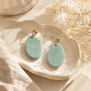 May include: A pair of light blue oval earrings with a textured pattern, displayed on a white ceramic dish. The earrings have gold-colored posts and are set against a neutral background with dried flowers.