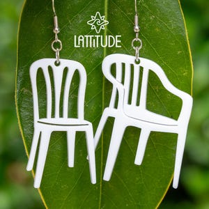 May include: White plastic chair earrings. The earrings are shaped like miniature versions of the classic white plastic lawn chair. They are suspended from silver-colored fish hook ear wires. The earrings are displayed on a green leaf with the word 'Lattitude' and a star logo in the background.