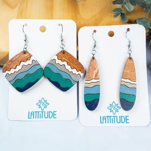 May include: Two pairs of wooden earrings with a wave design. One pair is diamond-shaped, the other is teardrop-shaped. The earrings feature brown, white, gray, teal, and navy blue colors. Each earring hangs from a silver-colored hook.