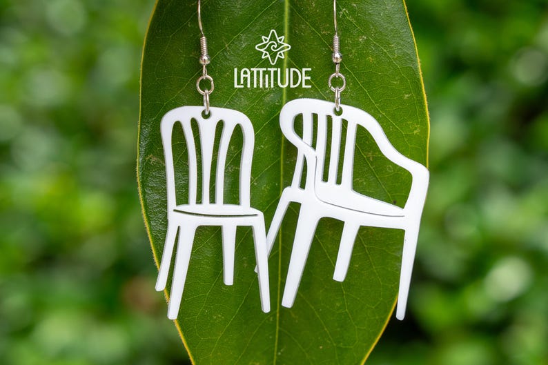 May include: White plastic chair earrings. The earrings are shaped like miniature versions of the classic white plastic lawn chair. They are suspended from silver-colored fish hook ear wires. The earrings are displayed on a green leaf with the word 'Lattitude' and a star logo in the background.