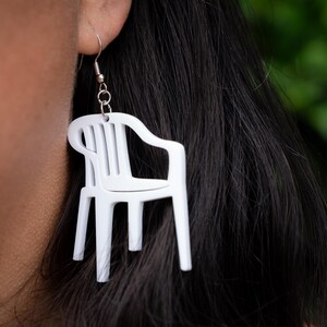 May include: White plastic chair earrings. The earrings are shaped like miniature lawn chairs, with a detailed design. The earrings are hanging from a silver hook. The background is a blurred green.