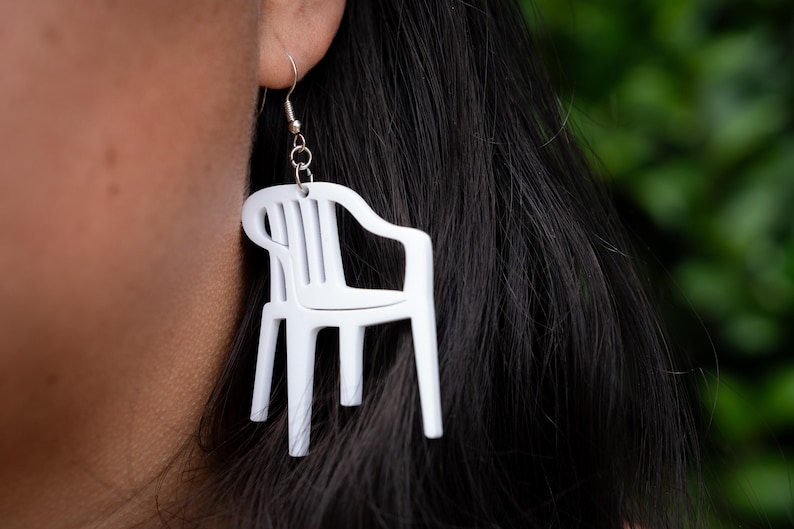 May include: White plastic chair earrings. The earrings are shaped like miniature lawn chairs, with a detailed design. The earrings are hanging from a silver hook. The background is a blurred green.