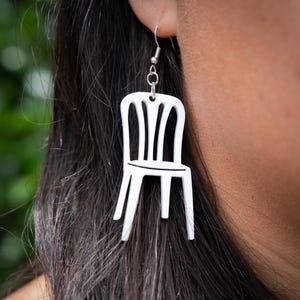 May include: White chair-shaped earrings. The earrings are a minimalist design, featuring a small, stylized chair silhouette. The chair has a back with vertical slats and a simple seat. The earrings hang from silver-colored ear wires.