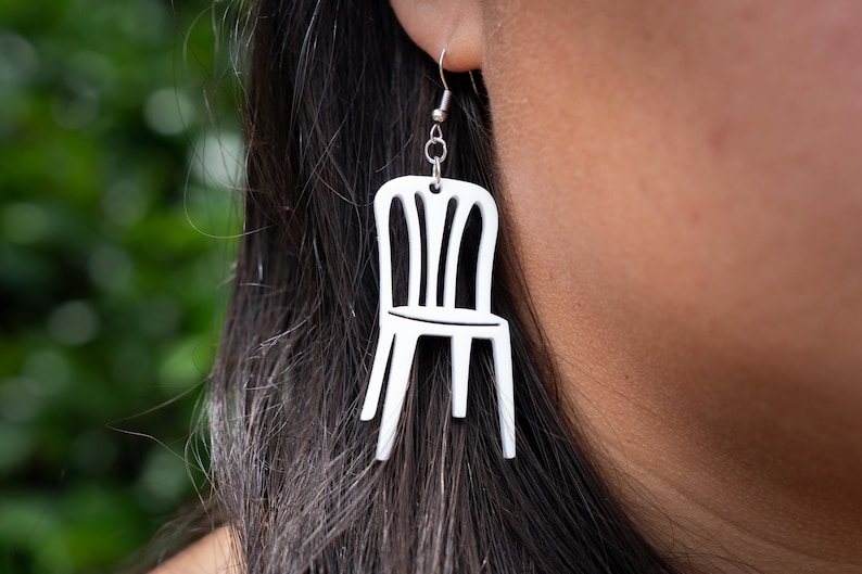 May include: White chair-shaped earrings. The earrings are a minimalist design, featuring a small, stylized chair silhouette. The chair has a back with vertical slats and a simple seat. The earrings hang from silver-colored ear wires.