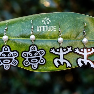 May include: Four pairs of earrings with silver hooks and white pearl beads. The earrings feature black and white designs, including stylized suns and human figures. The word "LATTITUDE" is printed on a green leaf.