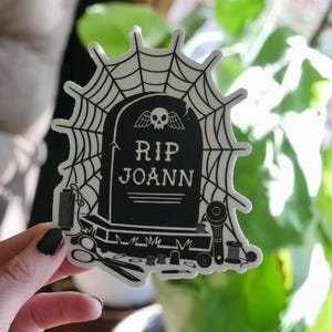 May include: A black and white sticker featuring a tombstone design with the text "RIP JOANN" and a skull. The sticker is surrounded by a spider web and sewing-related illustrations, including scissors and spools of thread.