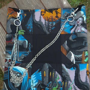 Puede incluir: Bolso cuadrado de patchwork con un centro de estrella negra y una correa de cadena plateada. El bolso presenta paneles con ilustraciones espeluznantes de vampiros, esqueletos y casas encantadas en tonos de azul, naranja y negro. El bolso está sobre una superficie de madera.
