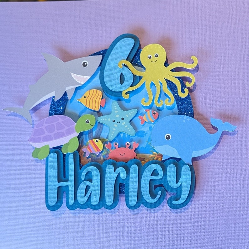 May include: A layered papercraft design featuring an underwater scene with a shark, turtle, whale, octopus, starfish, and fish. The number 6 and the name "Harley" are also included in the design. The color palette includes shades of blue, green, and orange.
