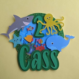 May include: A colorful, layered paper cake topper with an ocean theme. It features a gray shark, blue whale, yellow octopus, and various fish. The number "1" and the name "Cass" are also included, all set against a blue and green background.