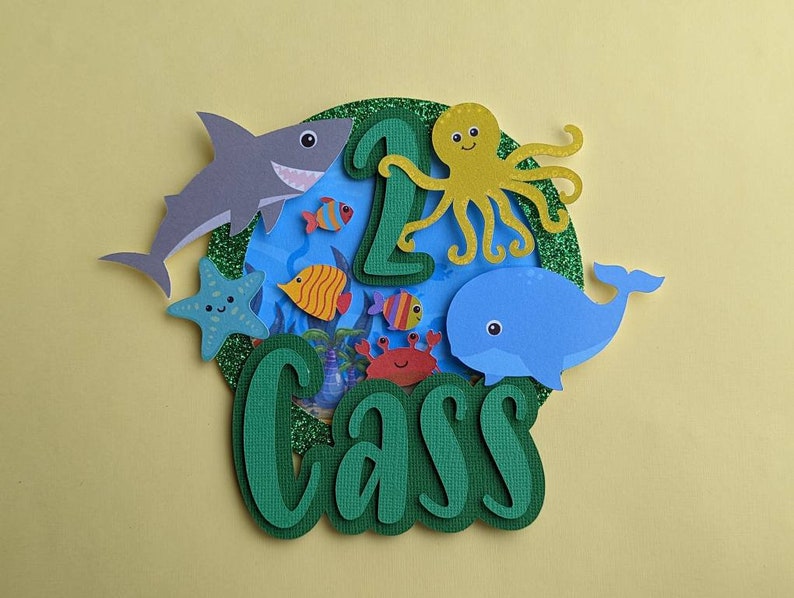 May include: A colorful, layered paper cake topper with an ocean theme. It features a gray shark, blue whale, yellow octopus, and various fish. The number "1" and the name "Cass" are also included, all set against a blue and green background.