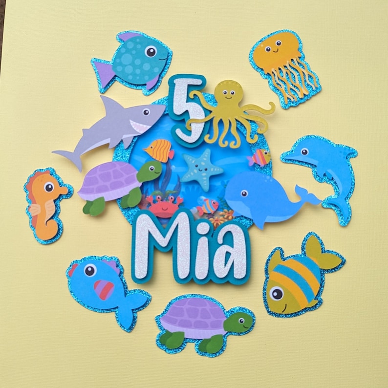May include: A colorful, layered cake topper with an ocean theme. It features various sea creatures like fish, a shark, a seahorse, and a whale, surrounding the number "5" and the name "Mia". The design includes glitter accents and a light blue and yellow color scheme.