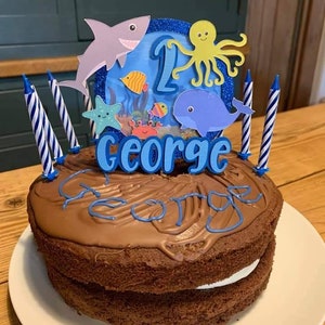 May include: A chocolate cake with a white filling, topped with chocolate frosting and blue lettering that spells "George". The cake is decorated with an ocean-themed topper featuring a shark, octopus, and whale, and has blue and white striped candles.