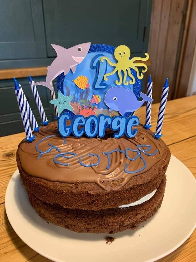 May include: A chocolate cake with a white filling, topped with chocolate frosting and blue lettering that spells "George". The cake is decorated with an ocean-themed topper featuring a shark, octopus, and whale, and has blue and white striped candles.