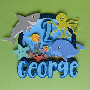 May include: A layered papercraft cake topper with an ocean theme. It features a gray shark, a yellow octopus, a blue whale, and various colorful fish. The number "1" and the name "GEORGE" are also included, all set against a blue and green background.