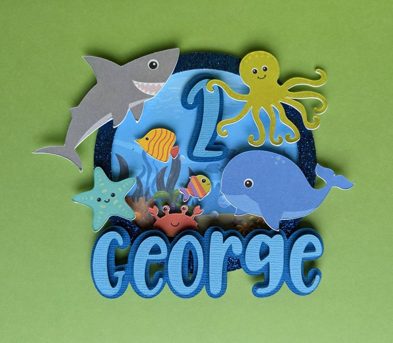 May include: A layered papercraft cake topper with an ocean theme. It features a gray shark, a yellow octopus, a blue whale, and various colorful fish. The number "1" and the name "GEORGE" are also included, all set against a blue and green background.