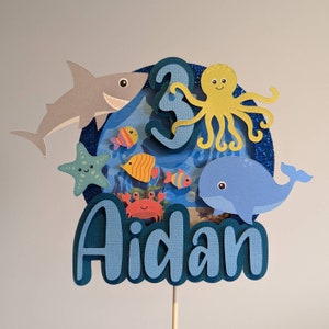 May include: A blue and white layered cake topper with an ocean theme. It features a shark, starfish, octopus, whale, crab, and fish. The number "3" and the name "Aidan" are also included.