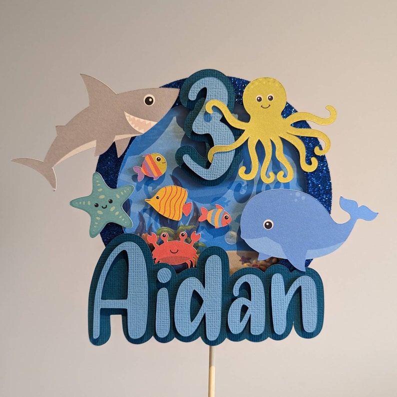May include: A blue and white layered cake topper with an ocean theme. It features a shark, starfish, octopus, whale, crab, and fish. The number "3" and the name "Aidan" are also included.