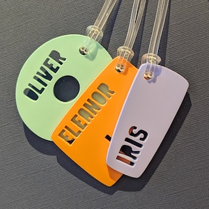 May include: Three luggage tags in green, orange, and lilac, each with a clear plastic loop. The tags have cut-out names: Oliver, Eleanor, and Iris. The tags are made of a smooth, solid material. Ideal for travel.