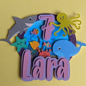 May include: A layered paper cake topper with an ocean theme. It features a gray shark, a blue whale, a green starfish, and various colorful fish. The number 7 and the name "Lara" are also included.