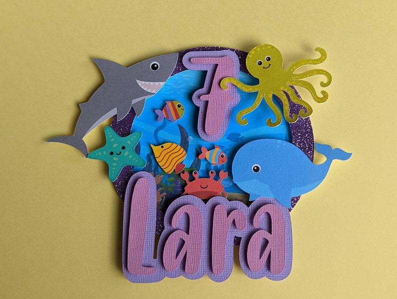 May include: A layered paper cake topper with an ocean theme. It features a gray shark, a blue whale, a green starfish, and various colorful fish. The number 7 and the name "Lara" are also included.