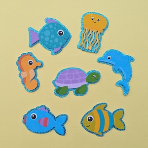 May include: A collection of colorful, glitter-edged sea creature cutouts, including a blue fish with purple accents, an orange seahorse, a yellow jellyfish, a blue dolphin, a purple turtle, and two additional fish.