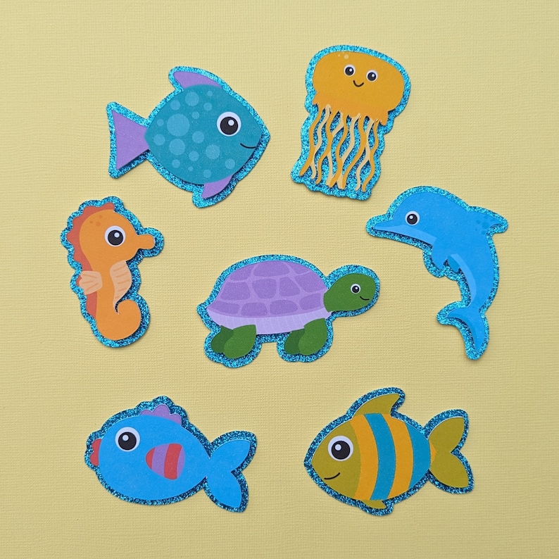 May include: A collection of colorful, glitter-edged sea creature cutouts, including a blue fish with purple accents, an orange seahorse, a yellow jellyfish, a blue dolphin, a purple turtle, and two additional fish.