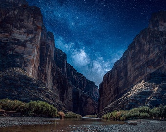 May include: A view of a canyon with a river running through it, under a dark blue sky filled with stars. The canyon walls are tall and rocky, and the river is calm and still.