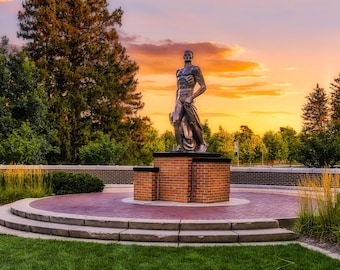 May include: A bronze statue of a man stands atop a brick and concrete pedestal. The statue is set against a backdrop of trees and a vibrant sunset. The statue is the focal point of a landscaped area.