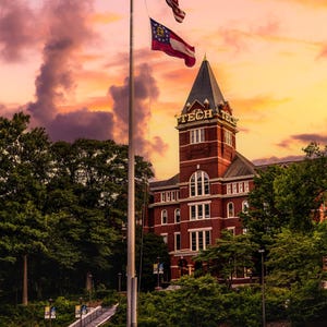 May include: A vibrant sunset over a brick building with a tall steeple. The American and Georgia state flags fly high. The building has the word "TECH" on its roof. Lush green trees and grass surround the building.