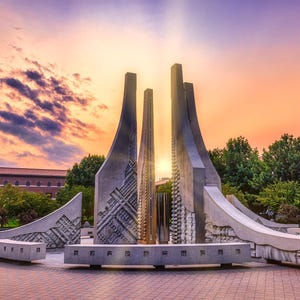May include: A large, abstract concrete sculpture with a central, vertical element and surrounding curved sections. The sculpture is set against a vibrant sunset sky with shades of orange, purple, and blue. Green trees and a brick building are in the background.
