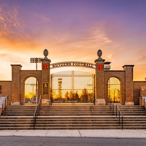 May include: A brick entrance to a football stadium with the words "RUTGERS FOOTBALL" above the gate. The sky is a gradient of orange and purple. The entrance has a set of stairs leading up to the gate.