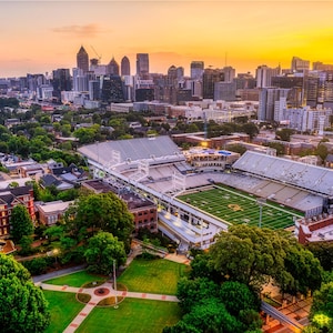 May include: Aerial view of a stadium with a green field, surrounded by buildings and lush green trees. The cityscape of Atlanta, Georgia, is visible in the background under a vibrant sunset.