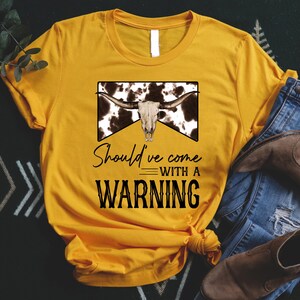 Könnte beinhalten: Ein gelbes T-Shirt mit einem schwarz-weißen Kuhschädel-Motiv und dem Text "Should've come with a warning".
