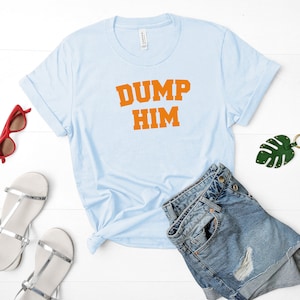 May include: Light blue t-shirt with the text "DUMP HIM" in orange letters. The shirt is laid flat on a white surface with a pair of silver sandals, a pair of sunglasses, and a green leaf keychain.