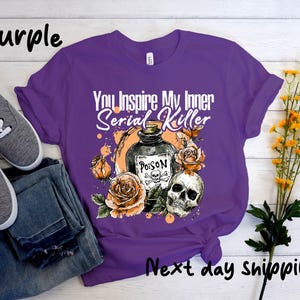 May include: Purple t-shirt with the text "You Inspire My Inner Serial Killer" and a graphic of a poison bottle, skull, and roses. The shirt is paired with gray shoes and blue jeans.