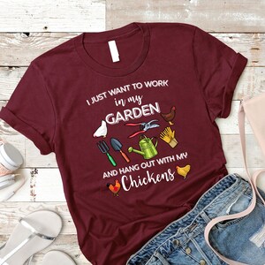 May include: Burgundy t-shirt with the text "I just want to work in my garden and hang out with my chickens". The shirt features a variety of gardening tools and chickens.