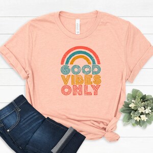 May include: Peach-colored t-shirt with a rainbow graphic and the text "GOOD VIBES ONLY" in orange, teal, and yellow. The shirt is paired with dark blue jeans. A small bouquet of greenery is in the background.