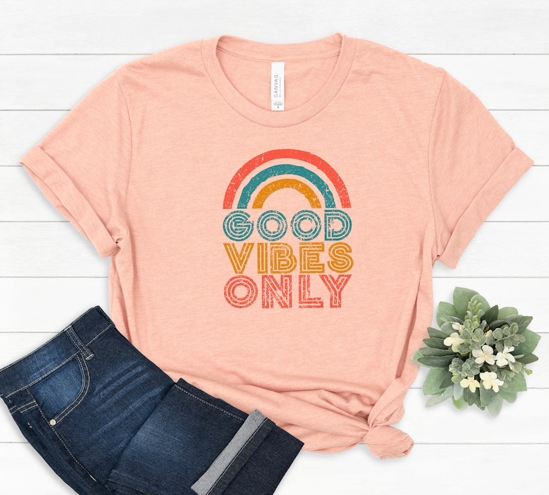 May include: Peach-colored t-shirt with a rainbow graphic and the text "GOOD VIBES ONLY" in orange, teal, and yellow. The shirt is paired with dark blue jeans. A small bouquet of greenery is in the background.