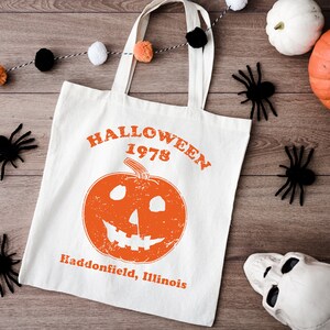 May include: A white canvas tote bag with black handles. The bag has an orange pumpkin with a jack-o'-lantern face and the text "HALLOWEEN 1978 Haddonfield, Illinois".