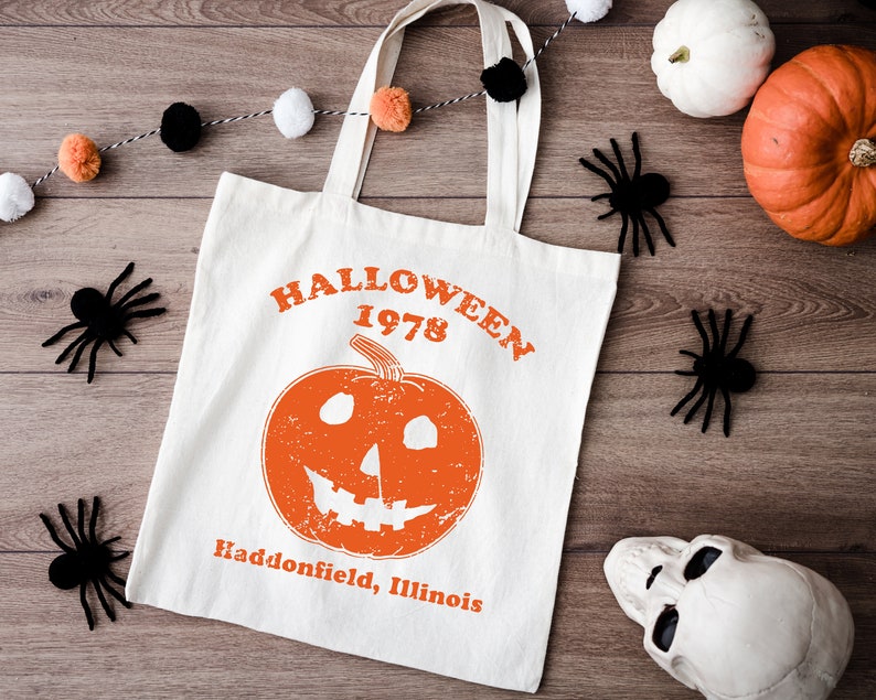 May include: A white canvas tote bag with black handles. The bag has an orange pumpkin with a jack-o'-lantern face and the text "HALLOWEEN 1978 Haddonfield, Illinois".