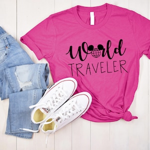 May include: A pink t-shirt with the text "World Traveler" in black script. The text includes a black globe and two black Mickey Mouse ears. The shirt is laid out on a white wooden surface with a pair of blue jeans and white sneakers.