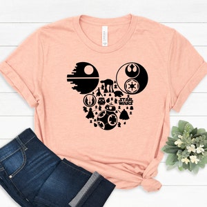 May include: A peach-colored t-shirt with a black silhouette of Mickey Mouse's head made up of Star Wars characters and symbols, including Darth Vader, R2-D2, Yoda, and the Death Star. The text "STAR WARS" is also included in the design.