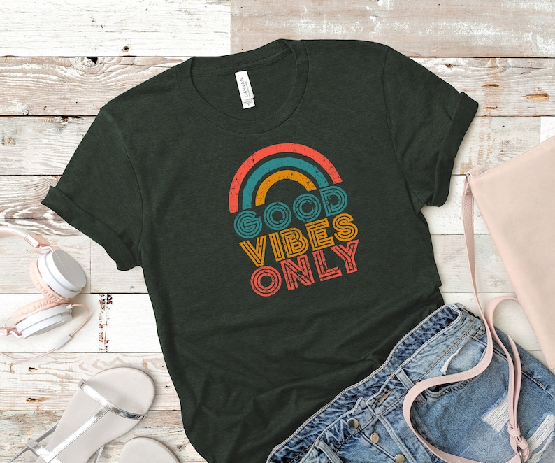 May include: Dark green t-shirt with a retro rainbow design and the words "GOOD VIBES ONLY" in orange, yellow, and teal. The shirt is folded, with a pair of denim shorts and a pink purse in the background.