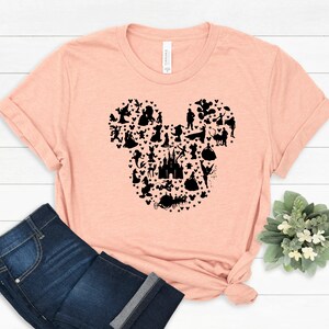 May include: A peach-colored t-shirt featuring a black silhouette of Mickey Mouse's head, filled with various Disney character silhouettes and stars. The shirt is paired with blue jeans.