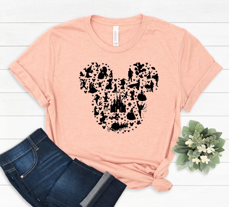 May include: A peach-colored t-shirt featuring a black silhouette of Mickey Mouse's head, filled with various Disney character silhouettes and stars. The shirt is paired with blue jeans.